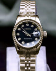 Gold watch with black face on a clear stand against a dark background