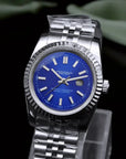 Silver watch with blue face on a clear stand against a dark background