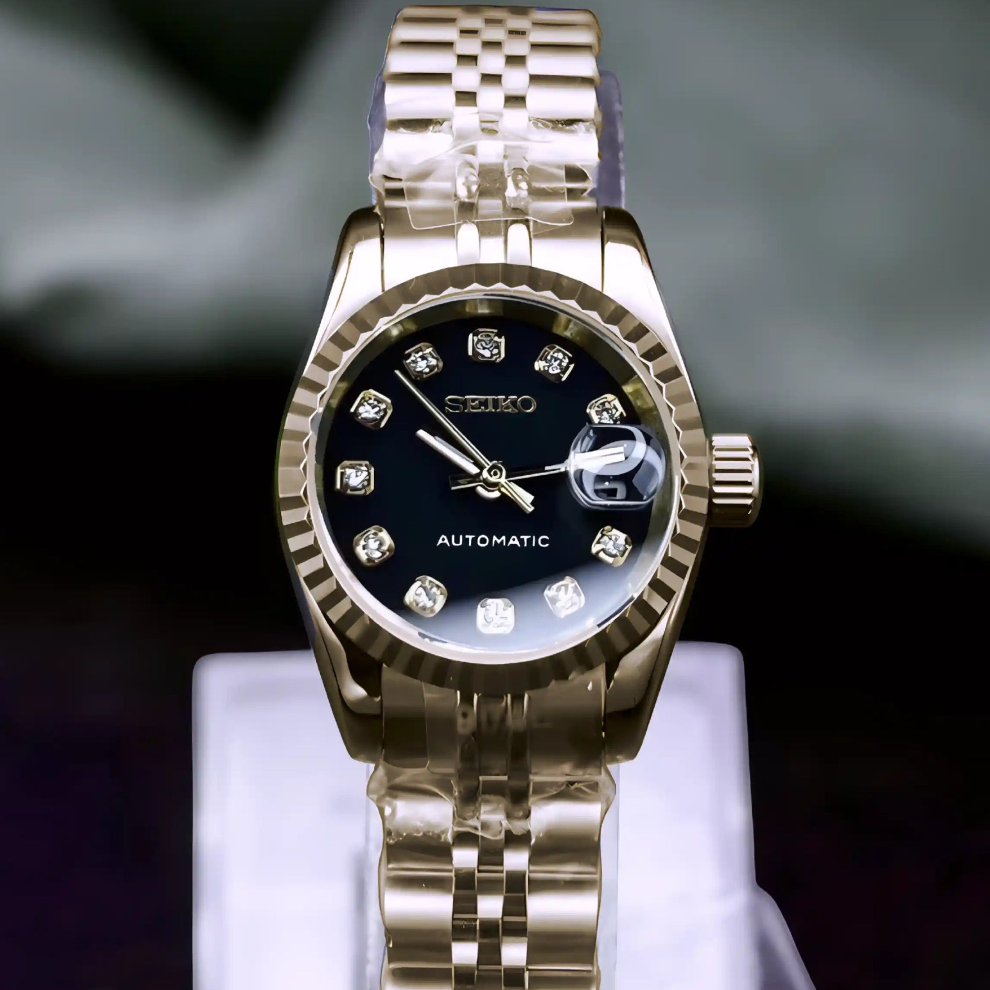 Gold watch with black face on a clear stand against a dark background