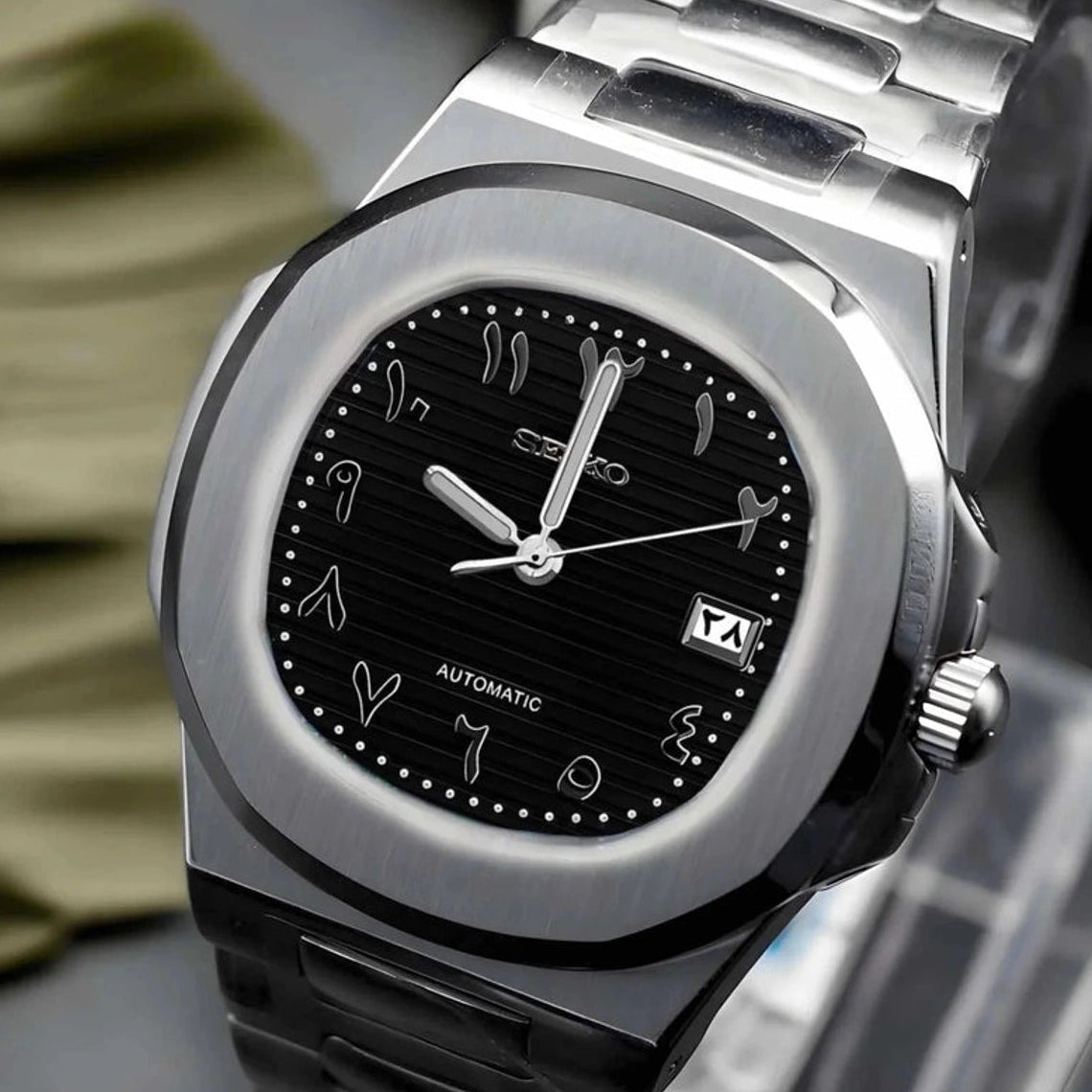 Silver watch with black arabic dial on a dark surface with green leaves in the background