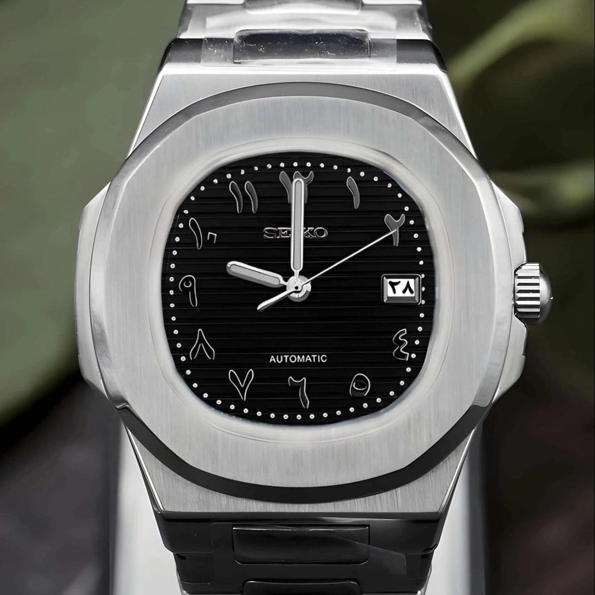 Silver watch with black arabic dial on a dark surface with green leaves in the background