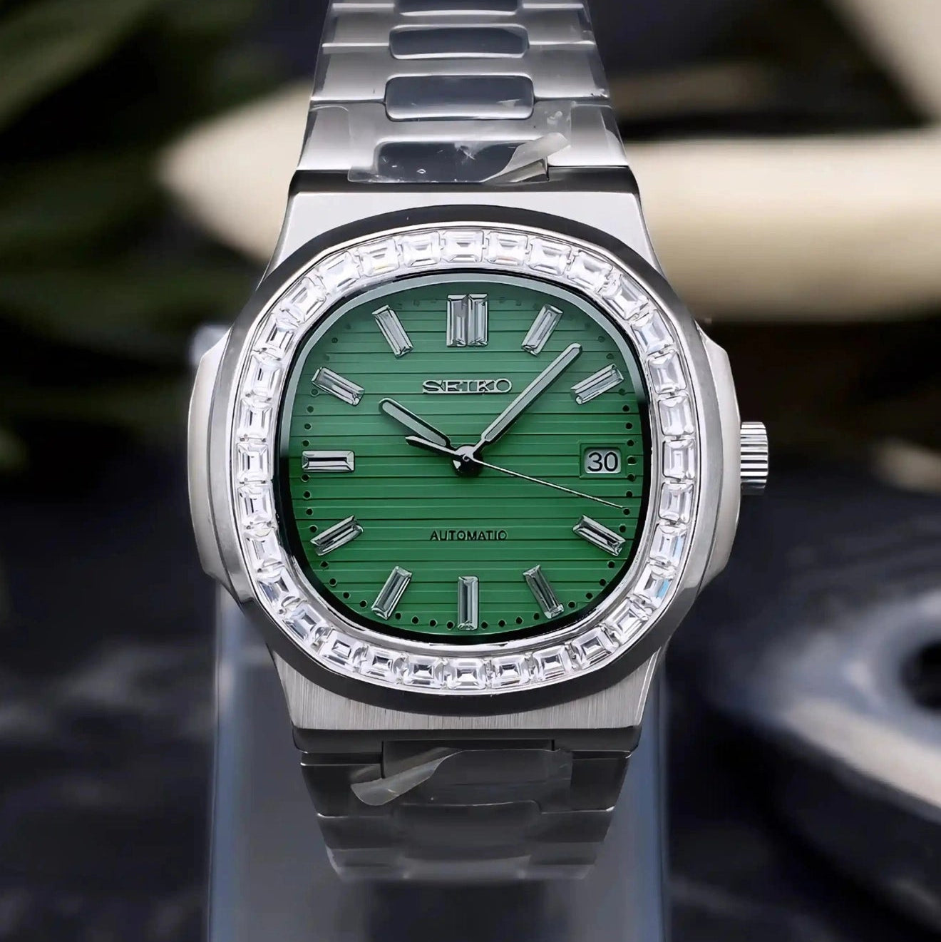 Silver watch with green face on a clear stand against a dark background
